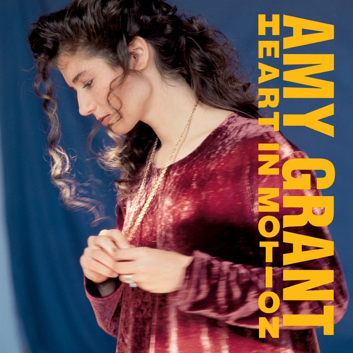 Album Art of Amy Grant's Heart In Motion album - In front a blue backdrop, a young white woman in a red dress is shown in profile and looking down and to the left. She's got a gold chain around her neck and her hands are clasping the item on the chain just in front of her chest. She's got long, brown curly hair. Printed above the photo on the right side is the artist's name down the right side vertically.</body></html>