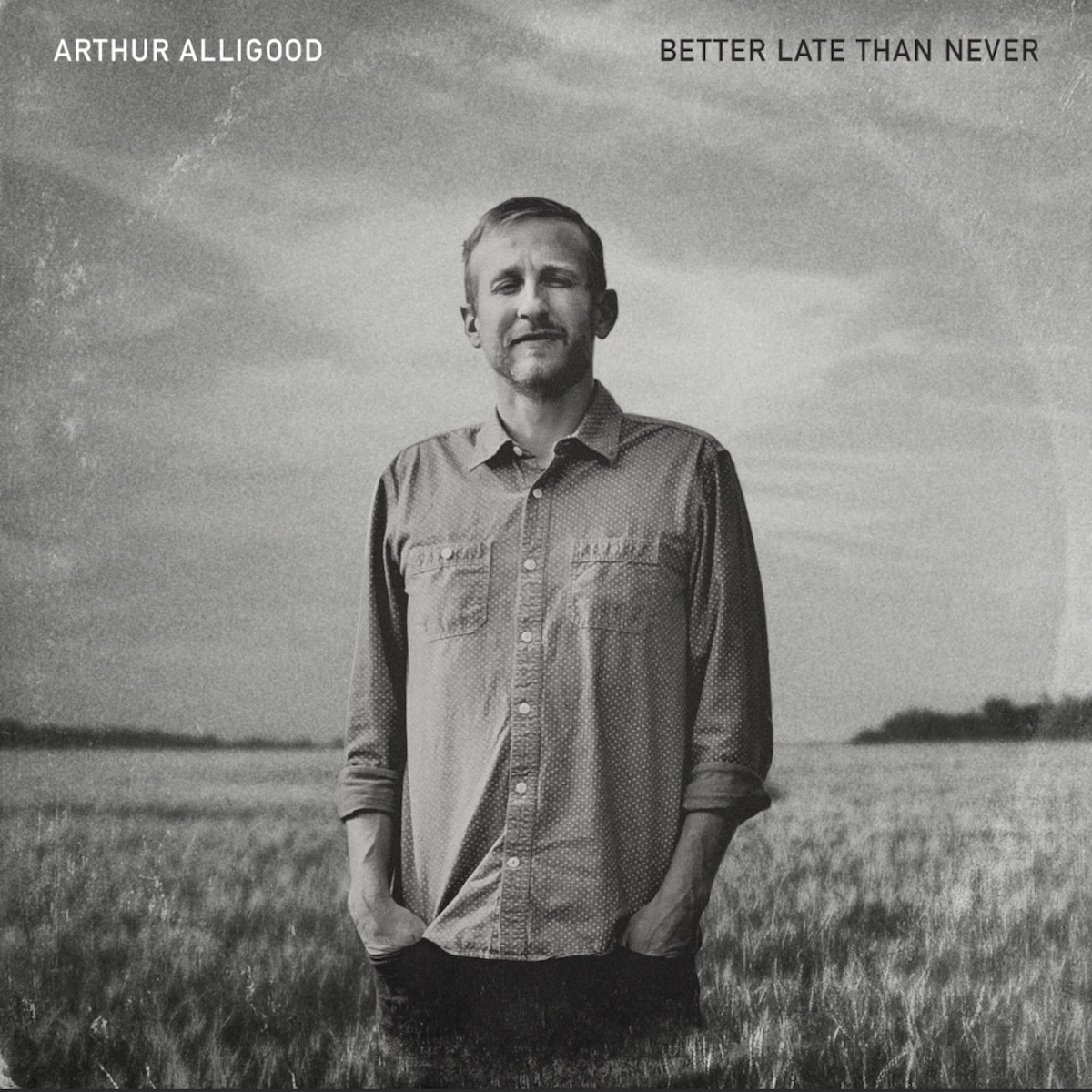 Album Art of Arthur Alligood's Better Late Than Never album - A black-and-white photo of a middle-aged white man standing with his hands in his pants pockets in a field of long grass. The field and sky are a gray color, and on the horizon some trees that are black. The man is pictured from the legs up, with dark pants and a light-colored collared shirt with pockets on both sides. He's god short hair and a short beard and mustache of a light color.</body></html>