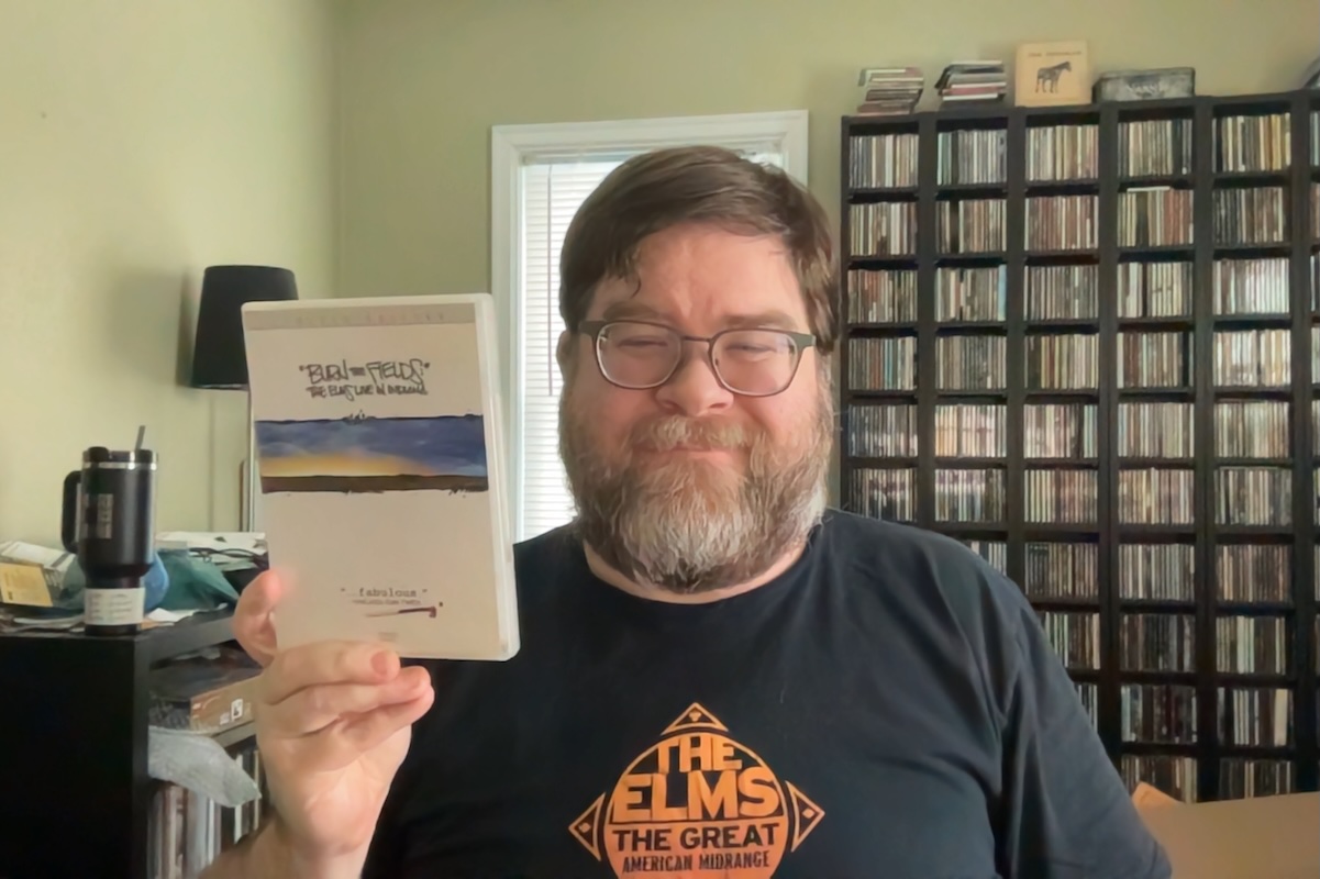 A photo of a man in a diffused sunlit room with light green walls holding a DVD of The Elms live concert film and smiling at the camera. He is also wearing a black shirt that says 'The Elms' on it in the center. Behind him in some CDs and vinyl, a lamp, and a water jug. He's a middle-aged white man with brown hair and a lighter-colored full beard and mustache.