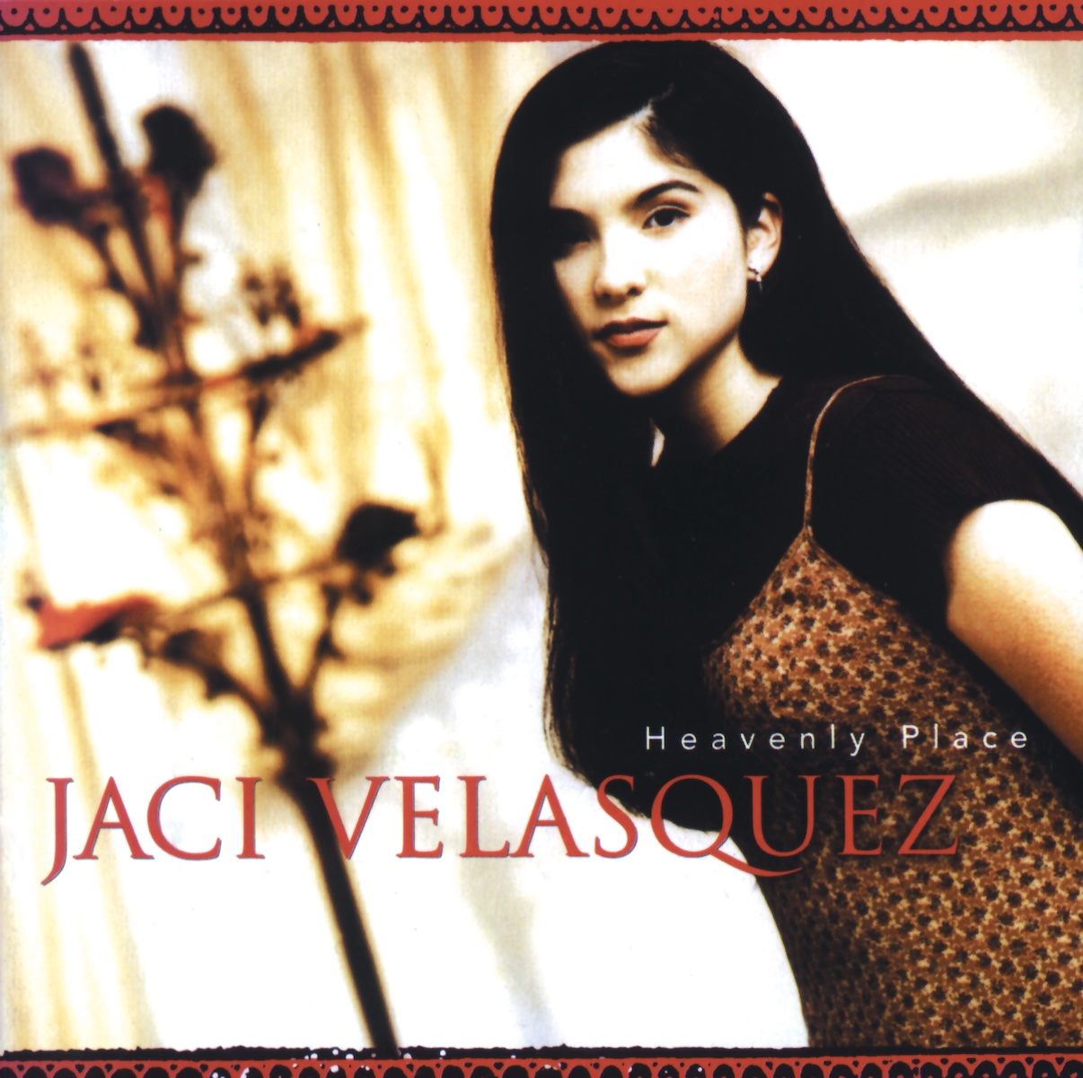 Album Art of Jaci Velasquez's Heavenly Place album - In a well-lit room, a photo of a teenage girl with long, black hair and a brown patterned dress is shown on the right side, with some sort of circular stand and curtains behind her on the left, though out of focus. The woman is looking at the camera and looks petite with a slight smile. Towards the bottom of the photo her name is printed in large red letters, with the album name in much smaller white letters above her name on the right. On the top and bottom there's a hand-drawn decorative border design in red and black.