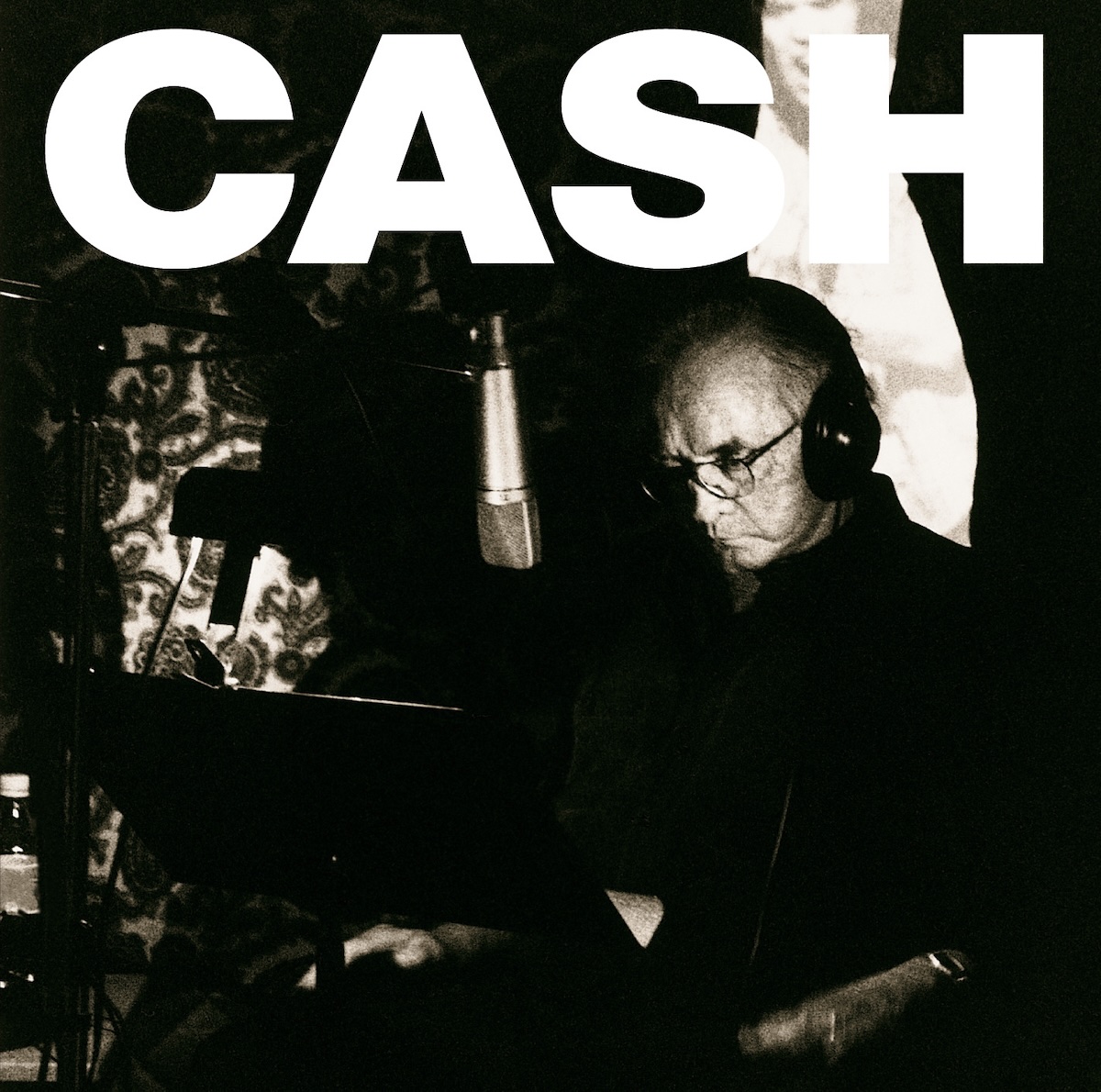 A Hundred Highways album - A black-and-white photo of an elderly Johnny Cash sitting in a darkened studio with a light shining on his face, sitting in front of a microphone and music stand. Most of the background is black, though there are some graphics pasted into the black in spots or maybe they are sitting on the back walls of the studio and other parts are obscured by something black. Across the top it just says 'Cash' in huge, white text.
