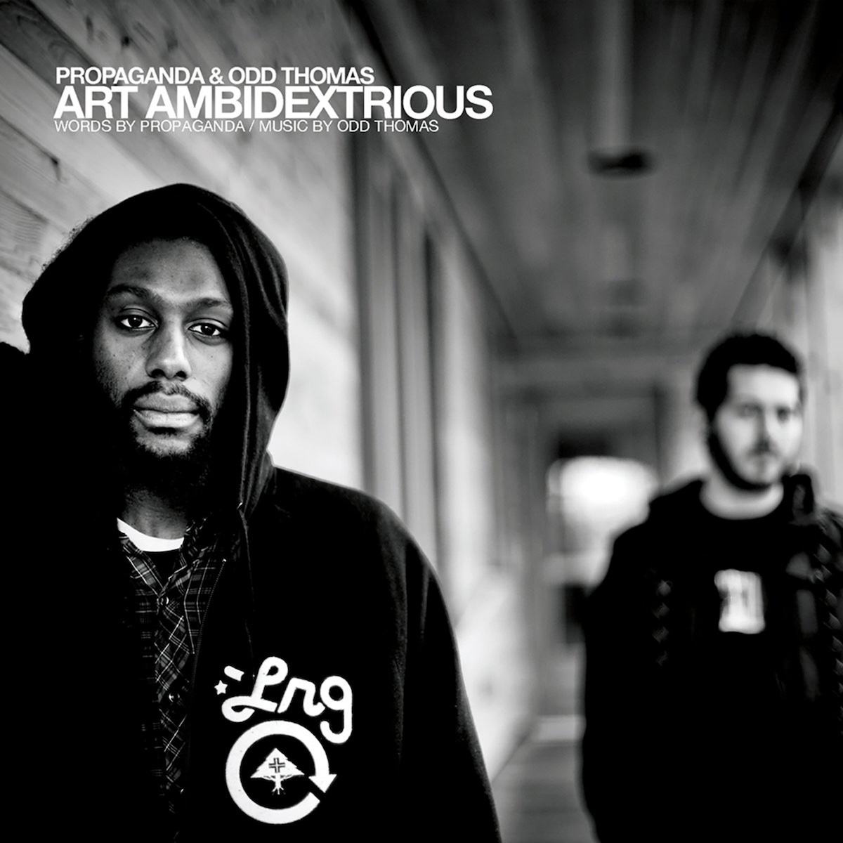 Album Art of Propaganda and Odd Thomas's Art Ambidextrous album - A black-and-white photo of two men in a long hallway. On the left is a black man in a black hoodie with the letters 'Lrg' printed on the front. A dozen feet down the hallway on the right is a white man with a beard and black hair and also in a black hoodie. Like most of the hallway in the background, this man in the background is a bit blurry. The hallway looks like it might have walls and a ceiling of wood of various shades.