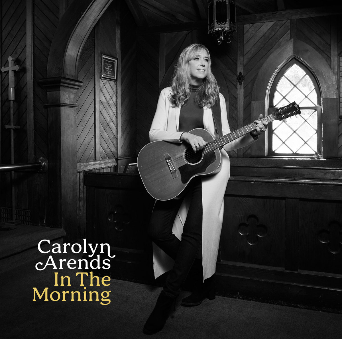Album Art of Carolyn Arends's In The Morning EP - A black-and-white photo of a white woman standing against a wooden railing and holding an acoustic guitar in some sort of small, rustic chapel with wooden walls and a small, simple glass window. She has blond hair down past her shoulders and is looking off to her left, while she has her hands on the guitar like she might start playing any moment.</body></html>