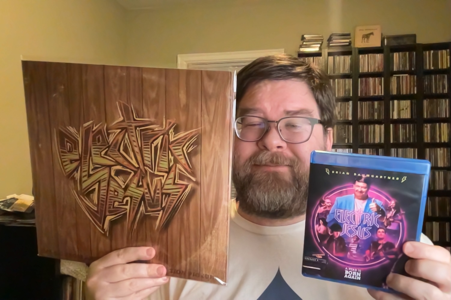 In a brightly lit office room with CD shelves behind, a rotund middle-aged white man holds a vinyl package of the Electric Jesus soundtracks as in one hand and the Electric Jesus movie Blu-Ray in another and is smiling to the camera between them.