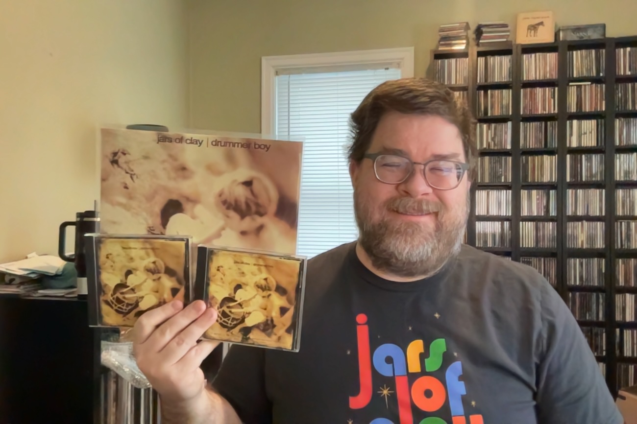 A photo of a rotund white man sitting in a room with shelves of CDs and records behind him. He's smiling and wearing a colorful 'Jars of Clay' T-shirt. He's holding a vinyl package of the EP as well as two CD versions in his right hand.