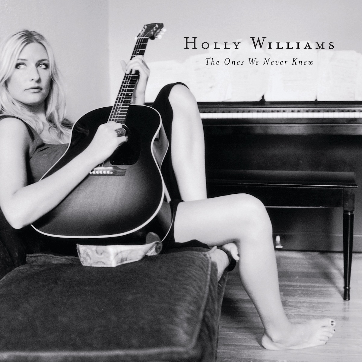 Album Art of Holly Williams's The Ones We Never Knew album - A black-and-white photo of a woman slouched on a couch with a large guitar on her lap, strumming it and looking to the left of the camera. The couch and her is mostly on the left side of the frame, with her right leg extending to the floor to the middle of the frame. The foot is on the wooden floor and behind her can be seen an upright piano with music spread out over the top of it.</body></html>