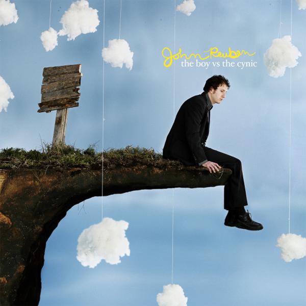 Album Art of John Reuben's The Boy Vs. The Cynic album - A photo of a white man with dark, curly hair sitting on the edge of a precipice and looking down with a bit of a blank stare. The precipice looks like it's a prop set up in a studio, as the dirt/stone of it sticks out comically far to where the person is sitting, like it was designed by a cartoonist. There's also a blank wooden sign next to the precipice but without any text on it.</body></html>