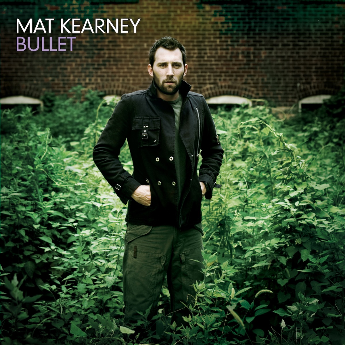 Album Art of Mat Kearney's Bullet album - A photo of a young man with a beard and short brown hair wearing green cargo pants and a green T-shirt with a black, double-breasted coat that is open slightly has his hands in the pockets of the coat. Behind him and around his feet is green foliage of some leafy plants, a bush or something. The plants are covering up the windows in a brick wall in the background.</body></html>