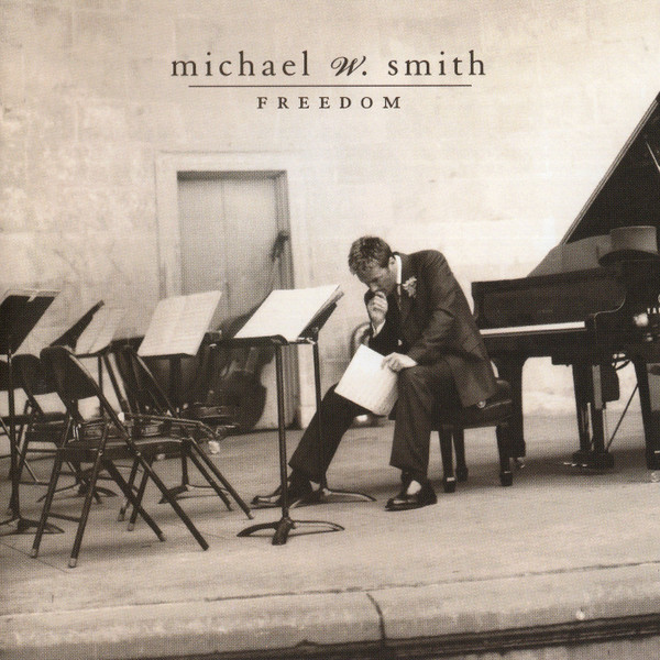 Album Art of Michael W. Smith's Freedom album - A black and white photo with a bit of a golden/brown sepia tone to it shows a lone man sitting on a stool in front of a grand piano, but facing away from the piano towards a small row of empty chairs with music stands and music sheets on the stands. The man is hunched over in contemplation holding the music sheets in his left hand, with his right hand up to his lips and looking down.</body></html>