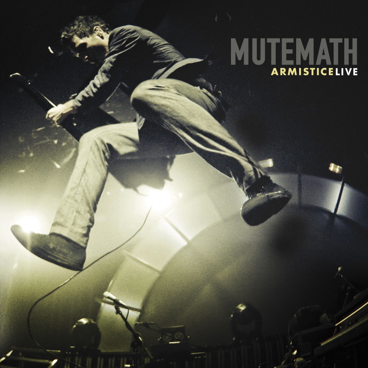 Album Art of Mutemath's Armistice Live album - A black-and-white photo where the white parts has a bit of a gold hue as well. The background is mostly dark and we can see that the picture is taken from the ground looking up. A man is jumping in the air and the closest thing to the camera are his feet. He's towering above the camera and also playing a keytar. Behind that, we can see the bright lights of a stage and an arched backdrop, with a few microphones and lights on the stage in front of it.</body></html>