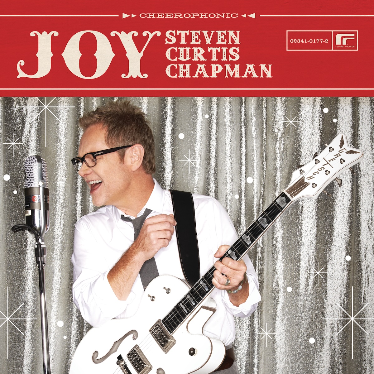 Album Art of Steven Curtis Chapman's Joy album - A color photo of a man in front of a grey and white background singing in profile to a silver microphone. He's wearing a white T-shirt and holding a white guitar with a black neck and black strap. He's singing open-mouthed with a smile and is wearing black-rimmed glasses. Above this photo, the top quarter or so is a red background with the word 'Joy' printed in huge letters, and the artist's name printed next to it on three different lines in smaller font. It also has the Reunion Records logo and at the top it says 'Cheerophonic' in small lettering.