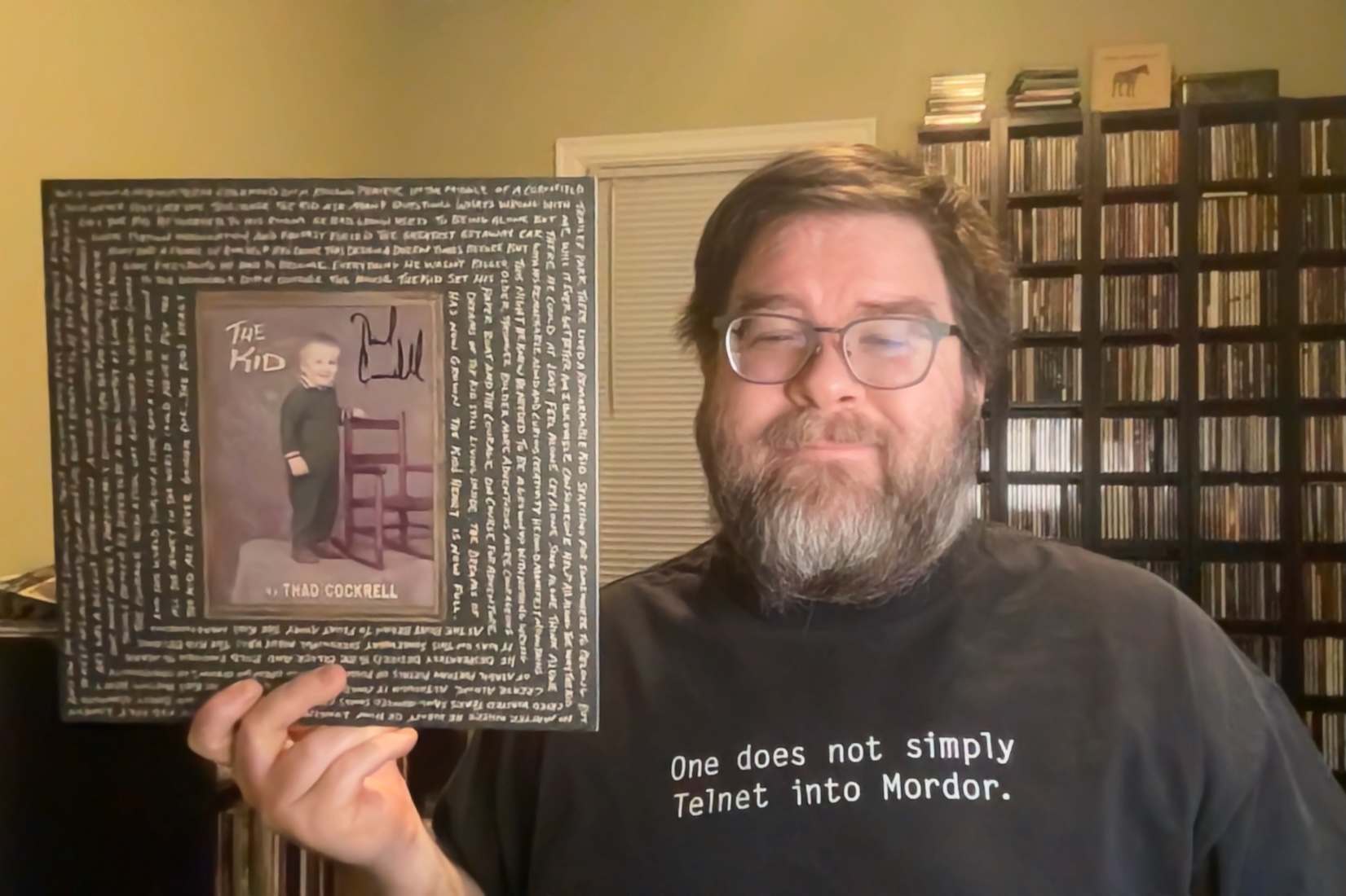 A middle-aged white man with a full beard and glasses is holding a copy of Thad Cockrell's The Kid album on vinyl next to his face. His T-shirt is black and says 'One must not Telnet into Mordor' in white typewriter font, and behind him is a window with the shade pulled down and a bunch of racks of CDs.