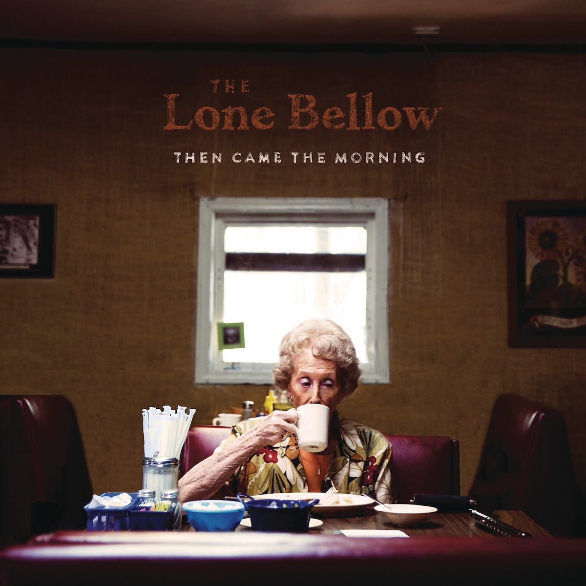 Album Art of The Lone Bellow's Then Came The Morning album - A photo of an older woman with white, permed hair sitting on the other end of a small table at a restaurant is sipping on a cup of coffee. She seems to be sitting by herself and have mostly finished a plate of food as it looks mostly empty. On the table are a couple empty side platters, a large jar with straws and a few other small bowls or trays with various condiments. Behind the woman is a brown wall with a small, square window. Up towards the ceiling on the wall is the band's name printed in an orange, with the album title printed in white in smaller text below it.