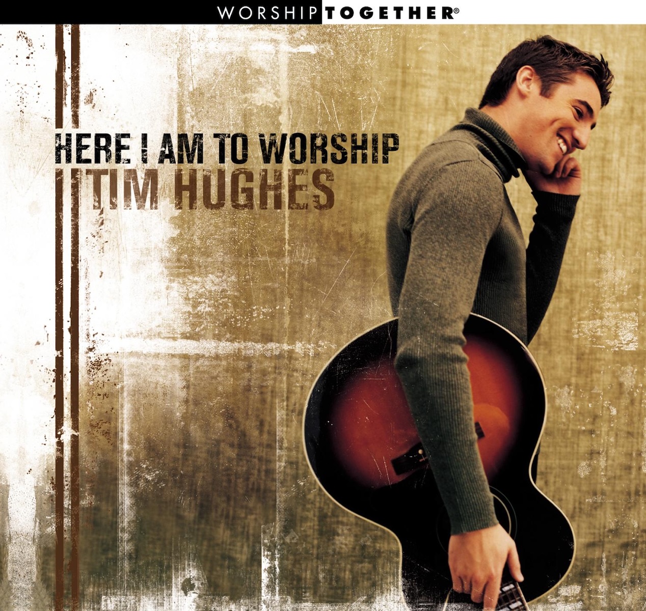 Album Art of Tim Hughes's Here I Am To Worship album - A young man with short brown hair and a long-sleeve grey turtleneck has an acoustic guitar on his right hip with the neck facing down, and he's smiling and looking down to the right of the frame. The background is brown mottled texture with some white paint thrown on top of it, and two vertical brown lines printed above that. The brown lines stop at the start of the album title, printed a bit above center in black block test, and below it the artist's name in a slightly larger brown.